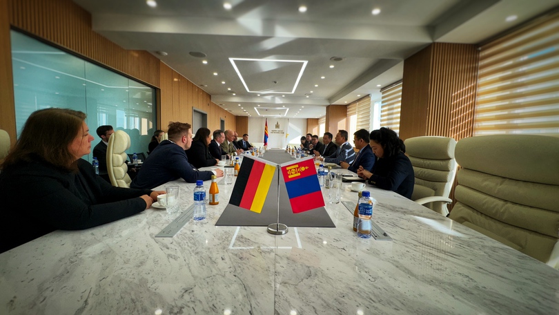Konferenztisch mit Teilnehmerinnen und Teilnehmern des Treffens. Im Vordergrund befindet sich ein Tischaufsteller mit den Flaggen von Deutschland und der Mongolei.